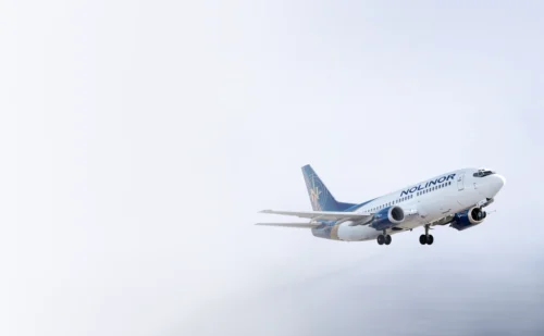 Un avion de la compagnie Nolinor en phase d'atterrissage, avec un ciel nuageux en arrière-plan.