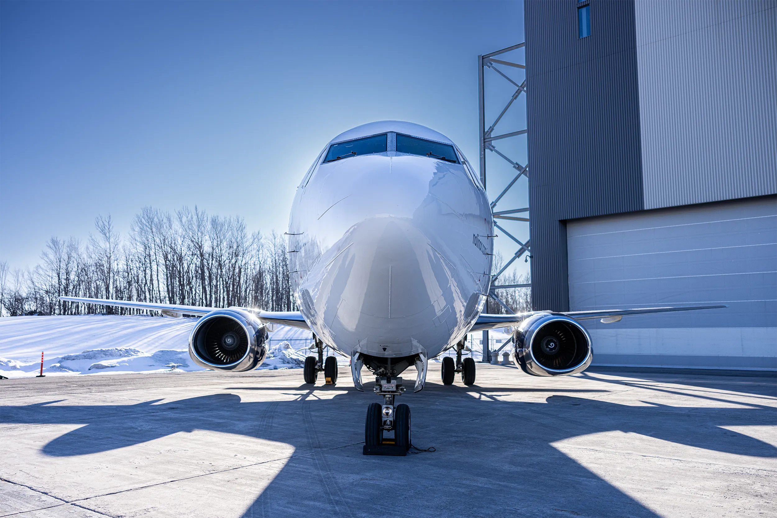Vue de face d’un avion 737-400 de Nolinor stationné devant un hangar, par temps ensoleillé avec de la neige au sol.