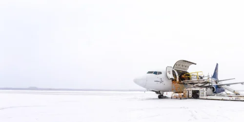 A plane is parked on a snowy runway, with a service staircase attached for boarding. Snow covers the ground, indicating winter weather.