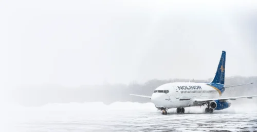 A Nolinor airplane on a snowy runway, surrounded by a winter landscape and falling snowflakes.
