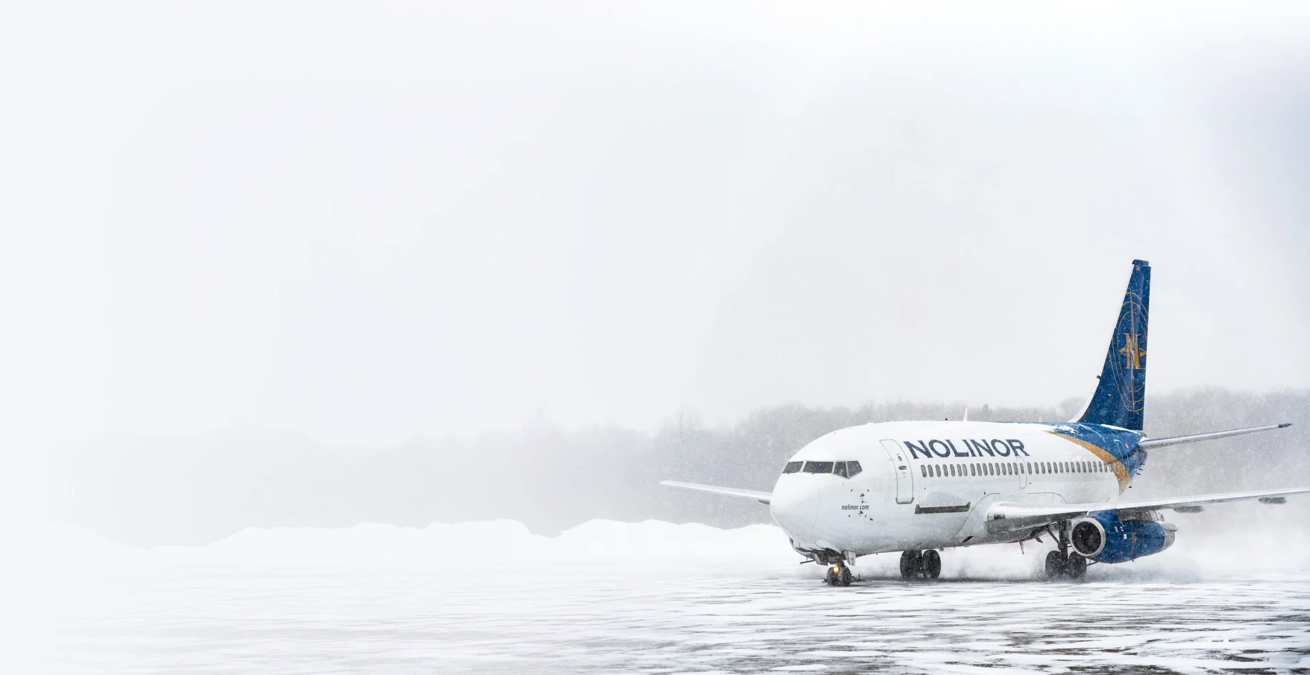 A Nolinor airplane on a snowy runway, surrounded by a winter landscape and falling snowflakes.