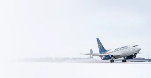 A Nolinor airplane takes off from a snowy runway, with a control tower visible in the background under overcast skies.