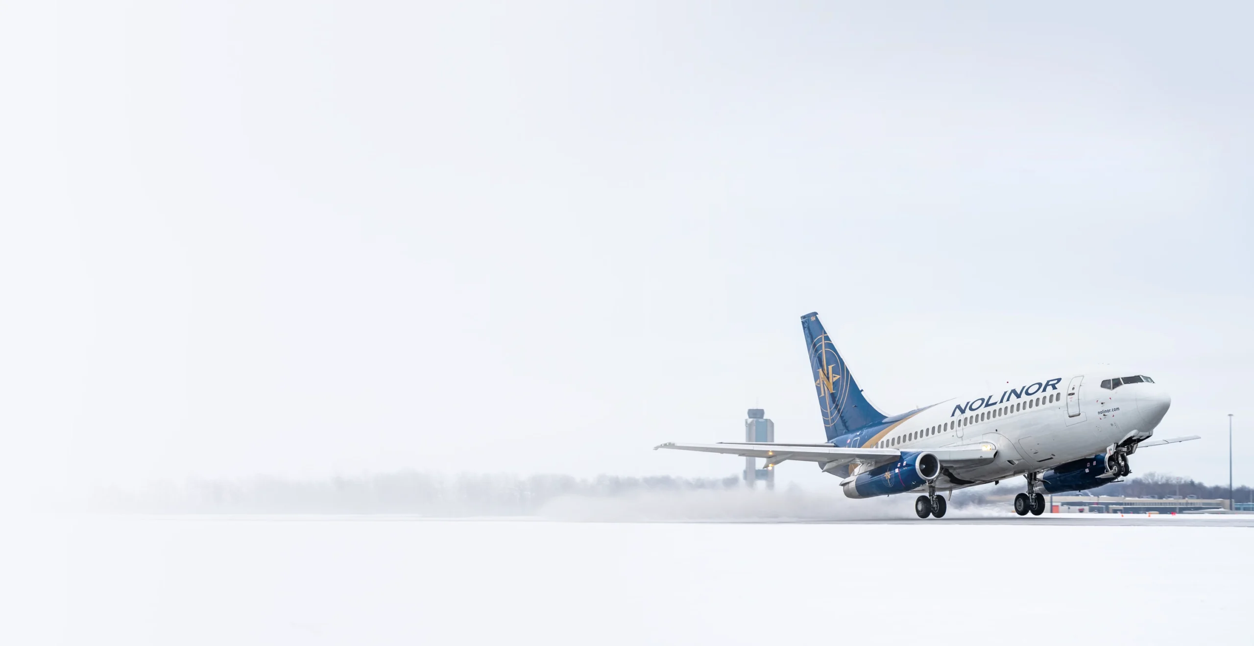 A Nolinor airplane takes off from a snowy runway, with a control tower visible in the background under overcast skies.