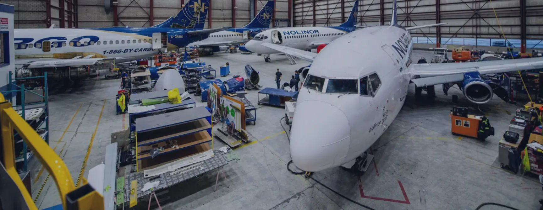 Airplanes in a hangar undergoing maintenance, with workers and equipment visible around them. Tools and parts are organized nearby.