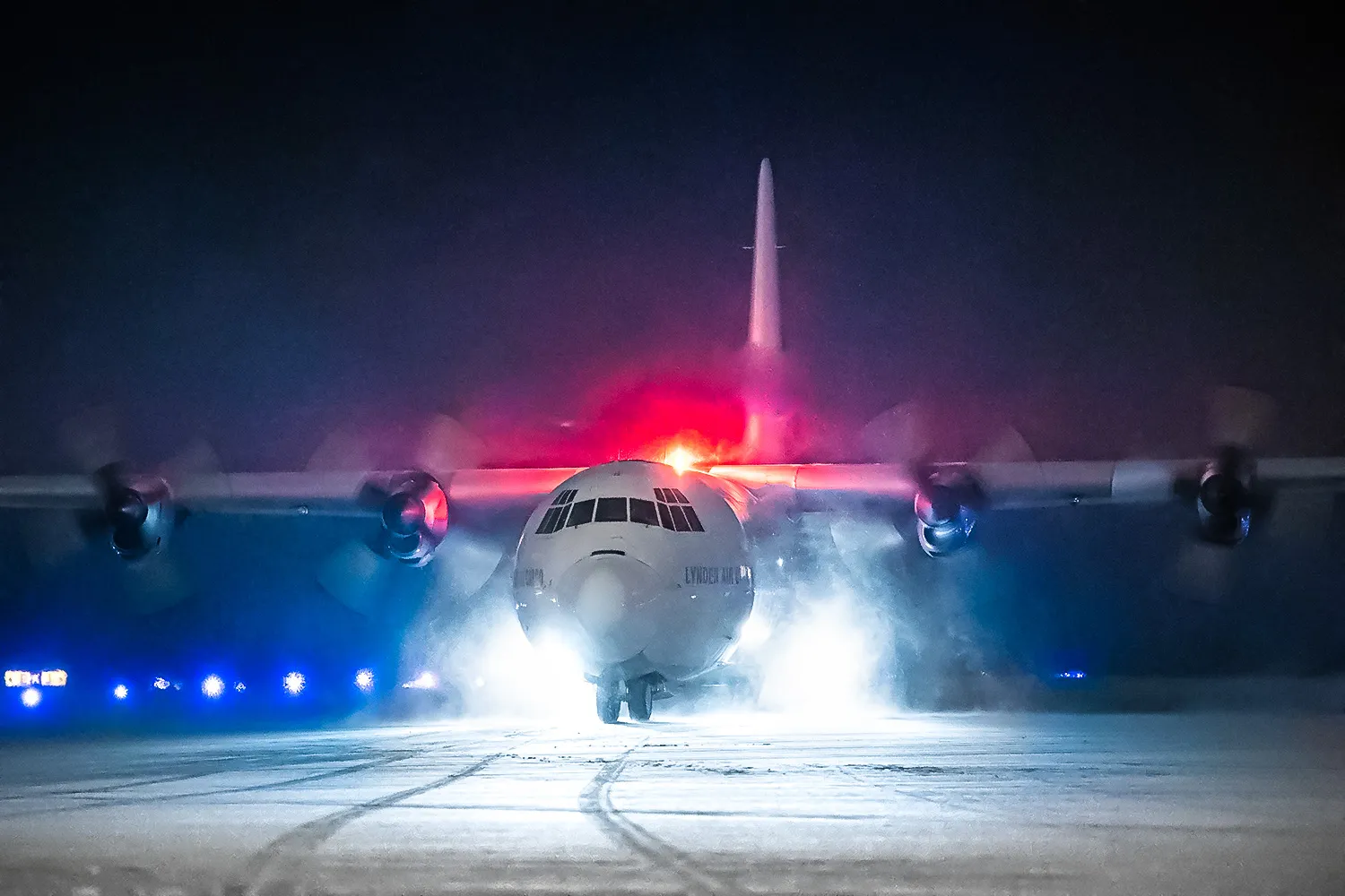 Un avion militaire se prépare à décoller sur une piste enneigée, illuminé par des feux clignotants dans la nuit.