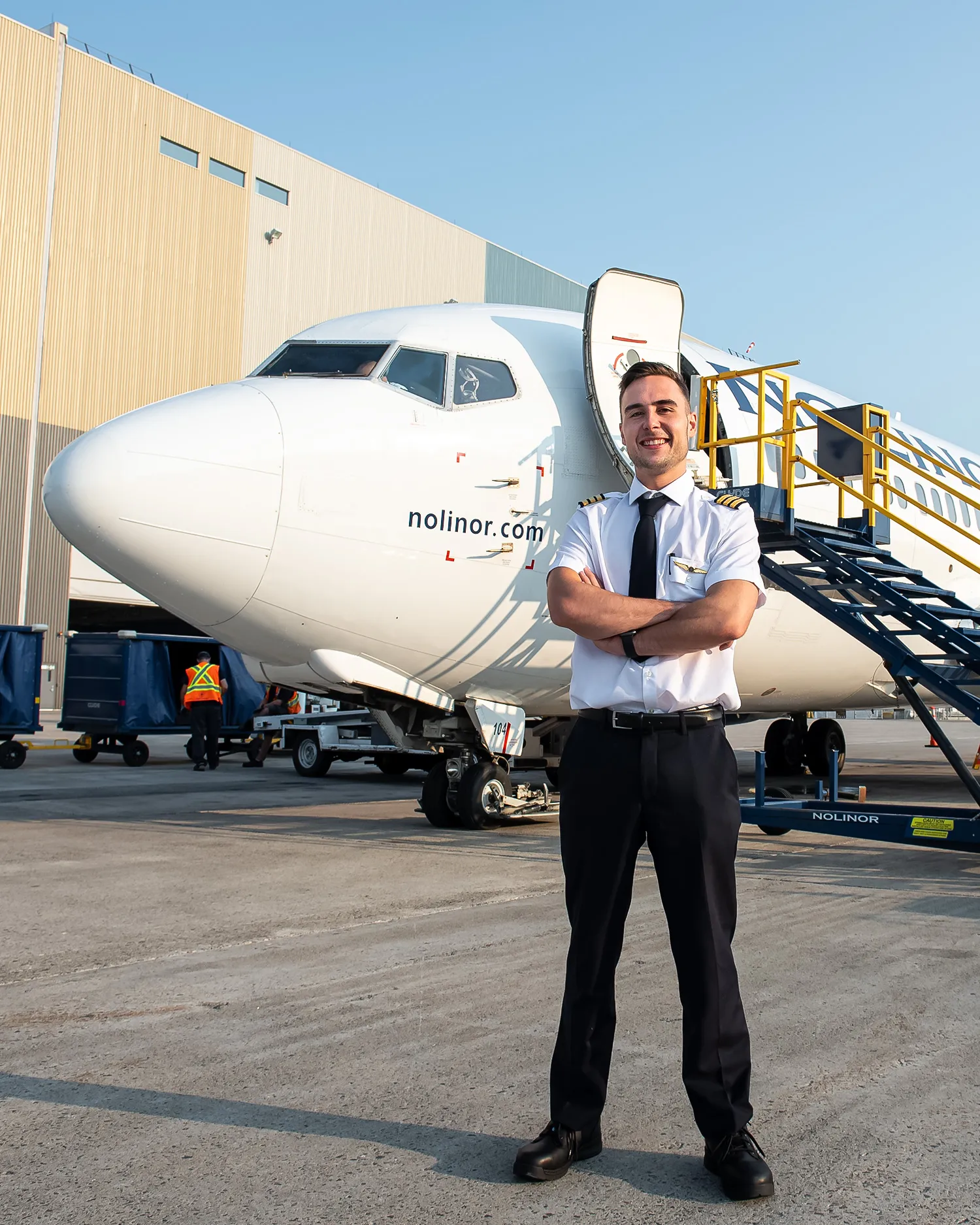 A pilot stands confidently in front of a Nolinor Airlines aircraft, with a boarding staircase nearby and airport staff in the background.