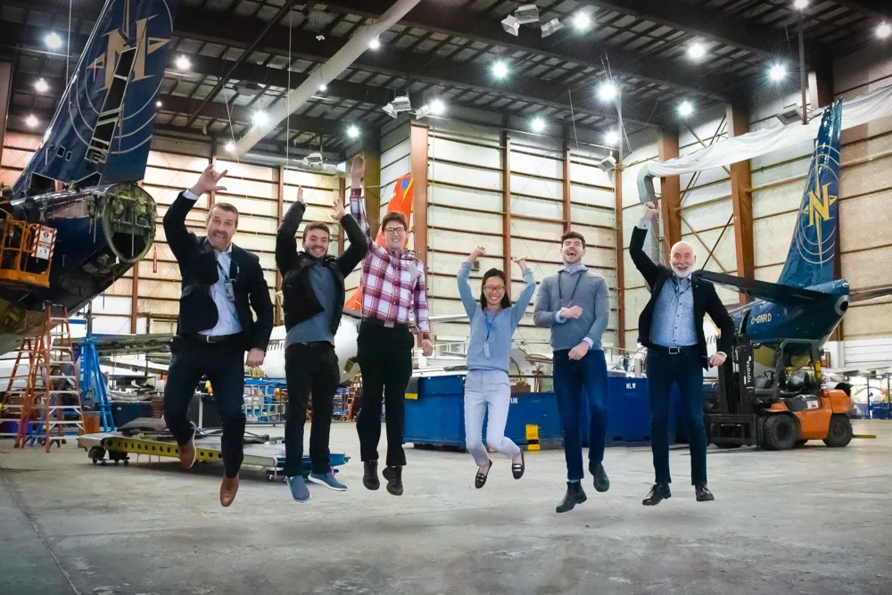 A group of six people in business attire jumps joyfully inside a hangar, with aircraft in the background.