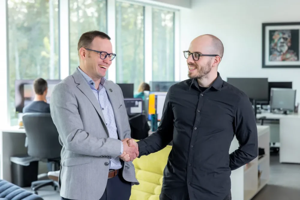 Two men shake hands in a modern office setting, smiling. In the background, colleagues work at computers.