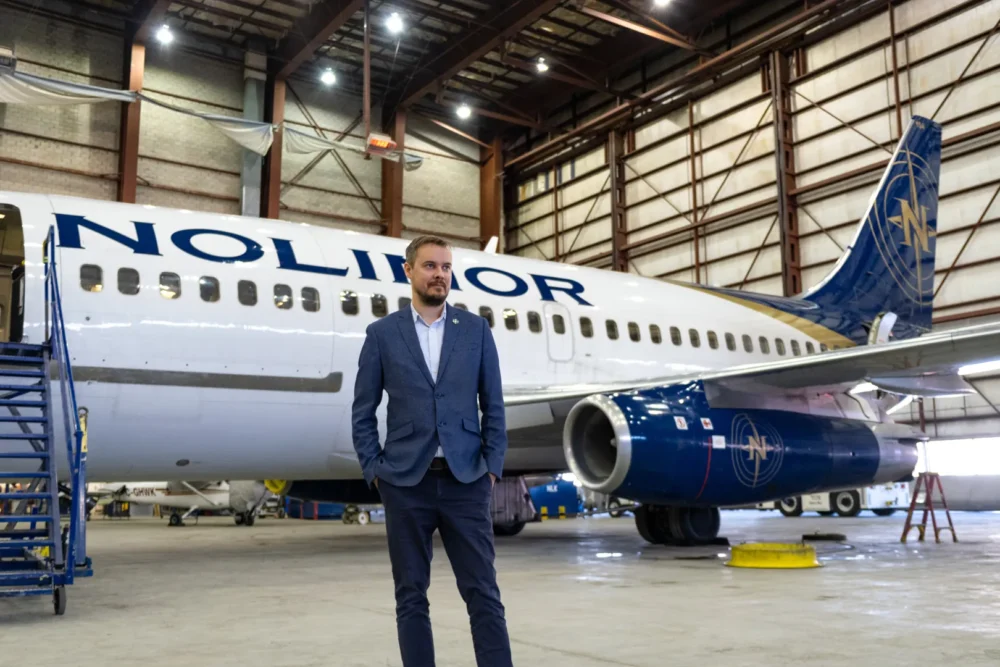 A man stands confidently in a hangar next to a blue and white aircraft marked with "Nolinor," showcasing the aviation environment.