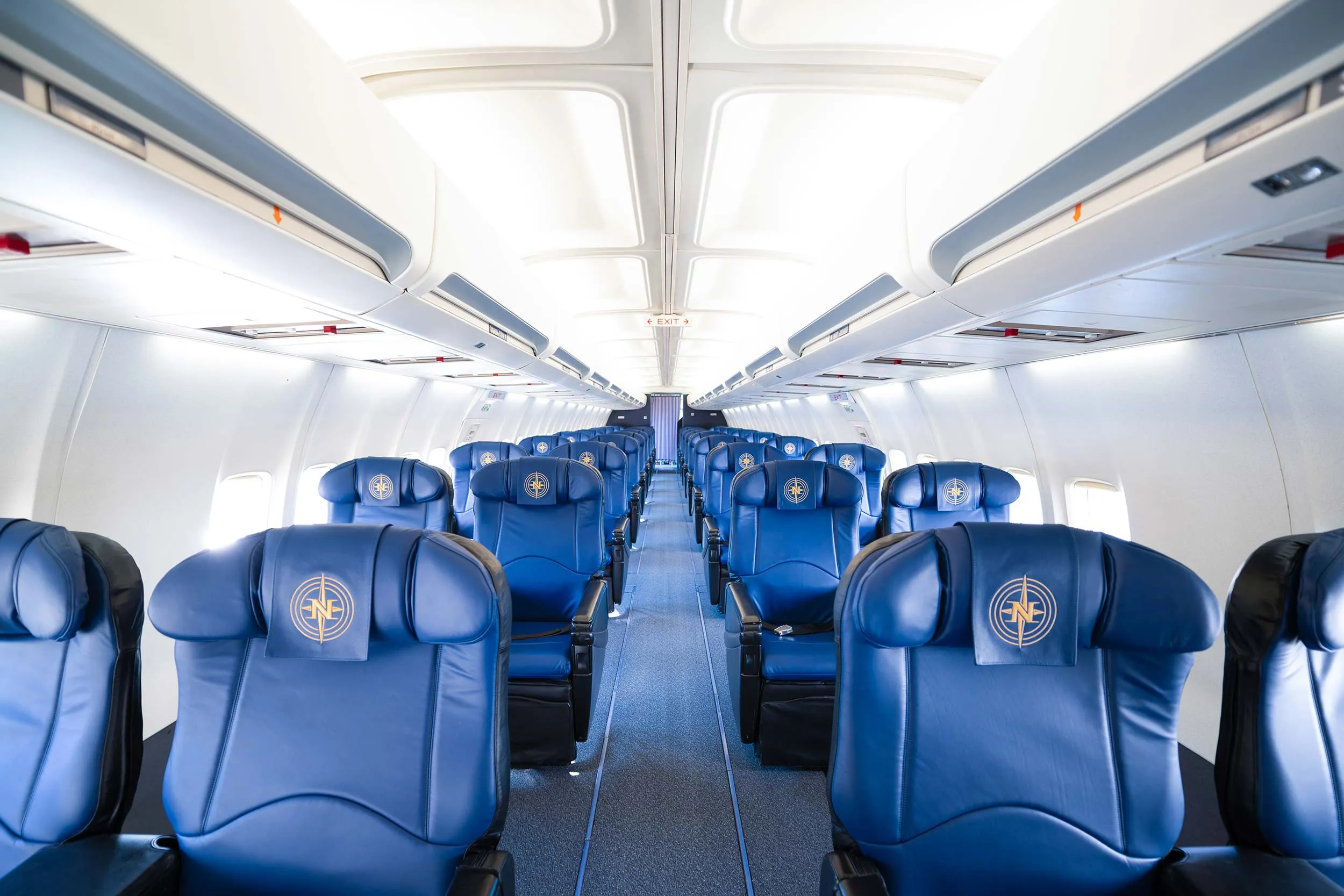 Interior of an airplane featuring rows of blue leather seats with a logo on the headrests, well-lit and spacious aisle.