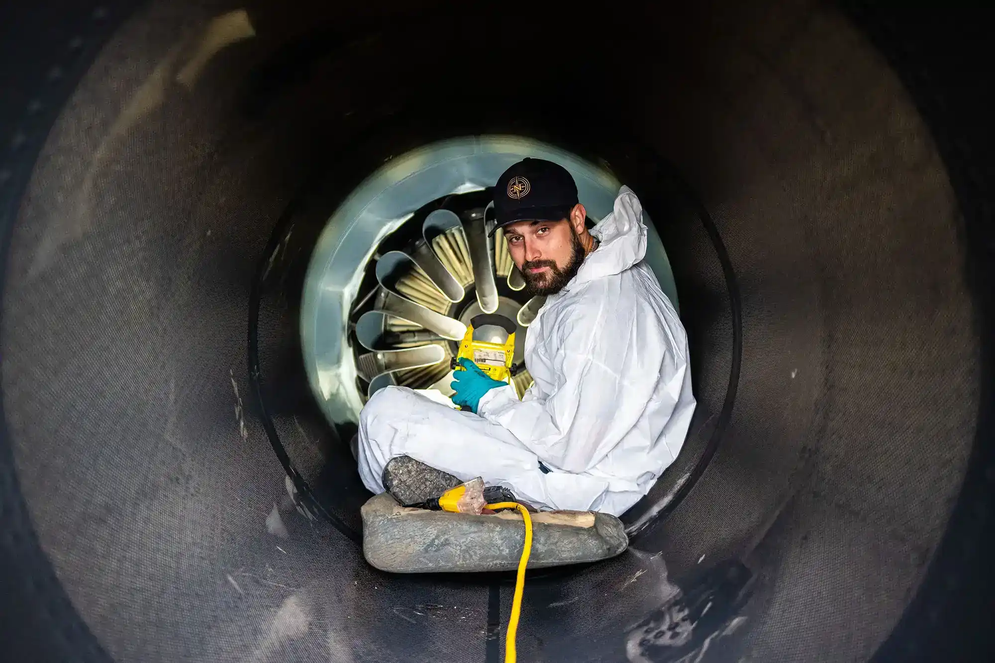 A man wearing a white jumpsuit and blue gloves is sitting inside a large black tube, holding a can of spray paint.