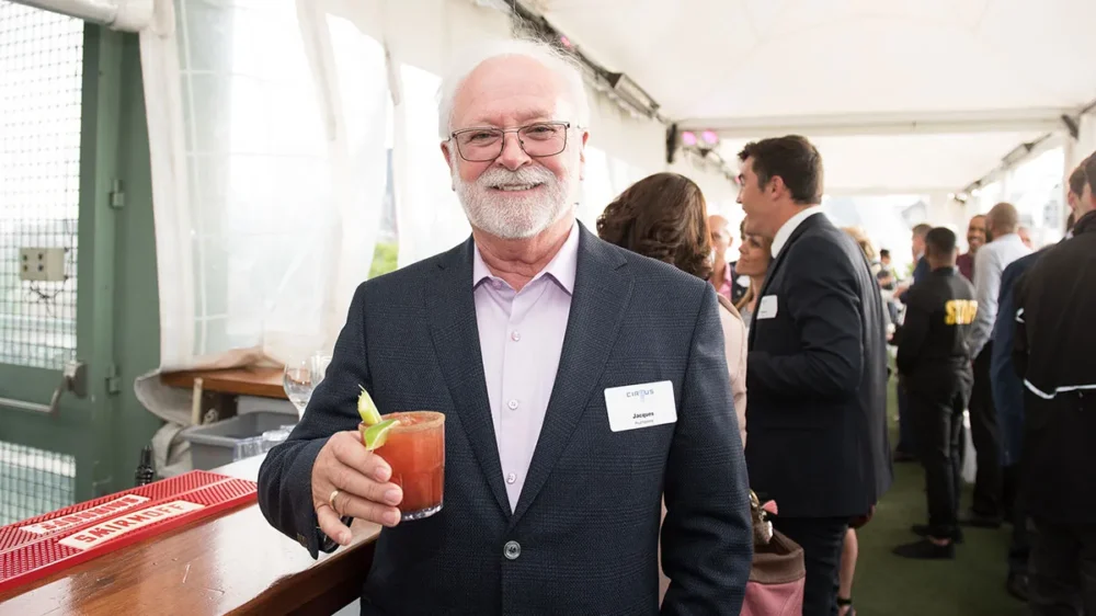 A smiling man in a suit holds a cocktail at a social event, with guests mingling in the background.