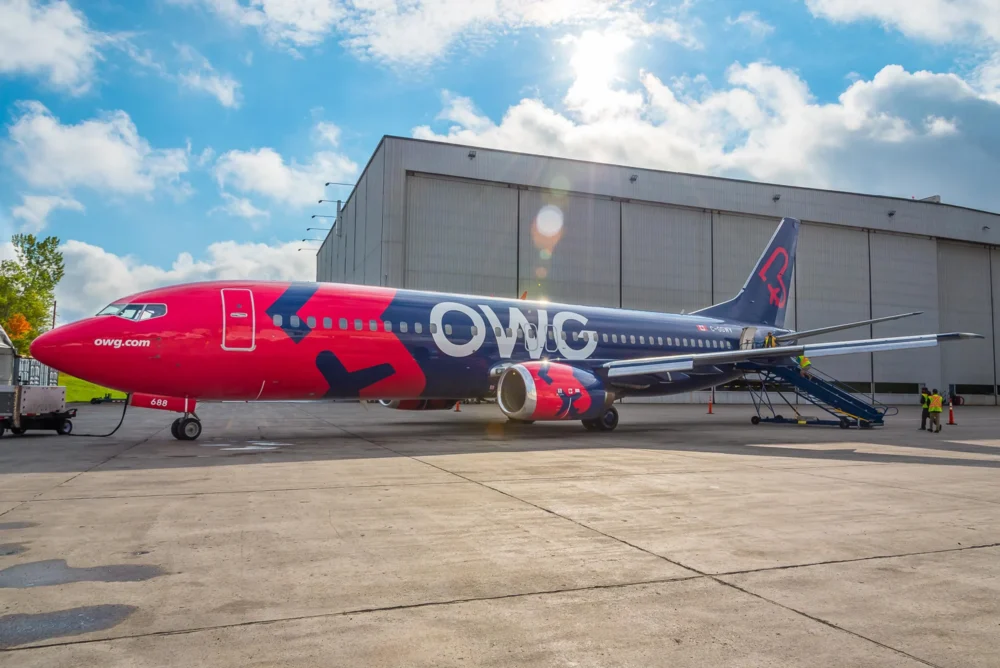 Un avion de ligne aux couleurs vives, rouge et bleu, est stationné devant un hangar. Une échelle est positionnée pour l'embarquement.