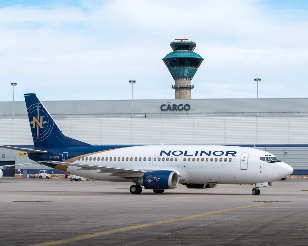 A Noliner cargo plane taxis on the runway near an airport control tower labeled "CARGO." The sky is partly cloudy.