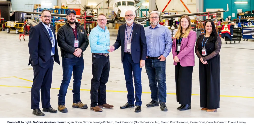 A group of seven professionals stands in an aircraft hangar, smiling and shaking hands, with a plane in the background.