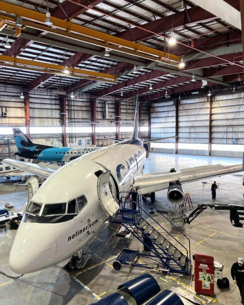 Un avion de ligne est stationné dans un hangar, avec des escaliers de service et des techniciens travaillant à l'intérieur. D'autres avions sont visibles en arrière-plan.
