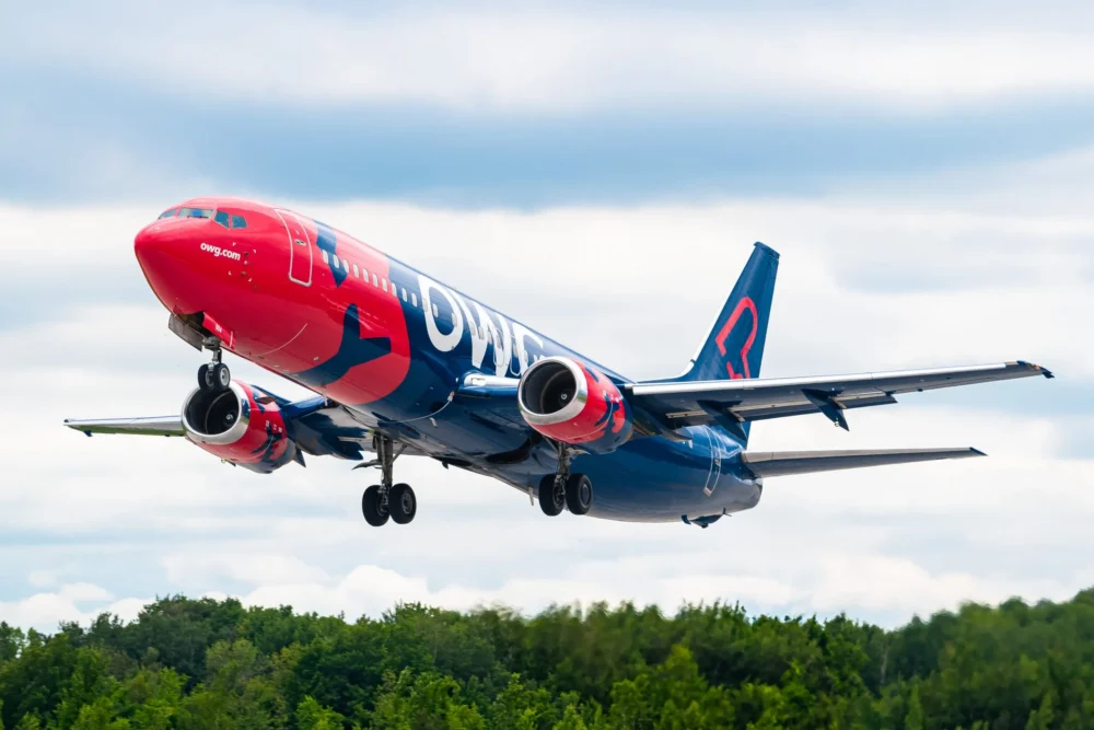 A brightly colored airplane with a red and blue design is taking off against a cloudy sky, surrounded by green trees.