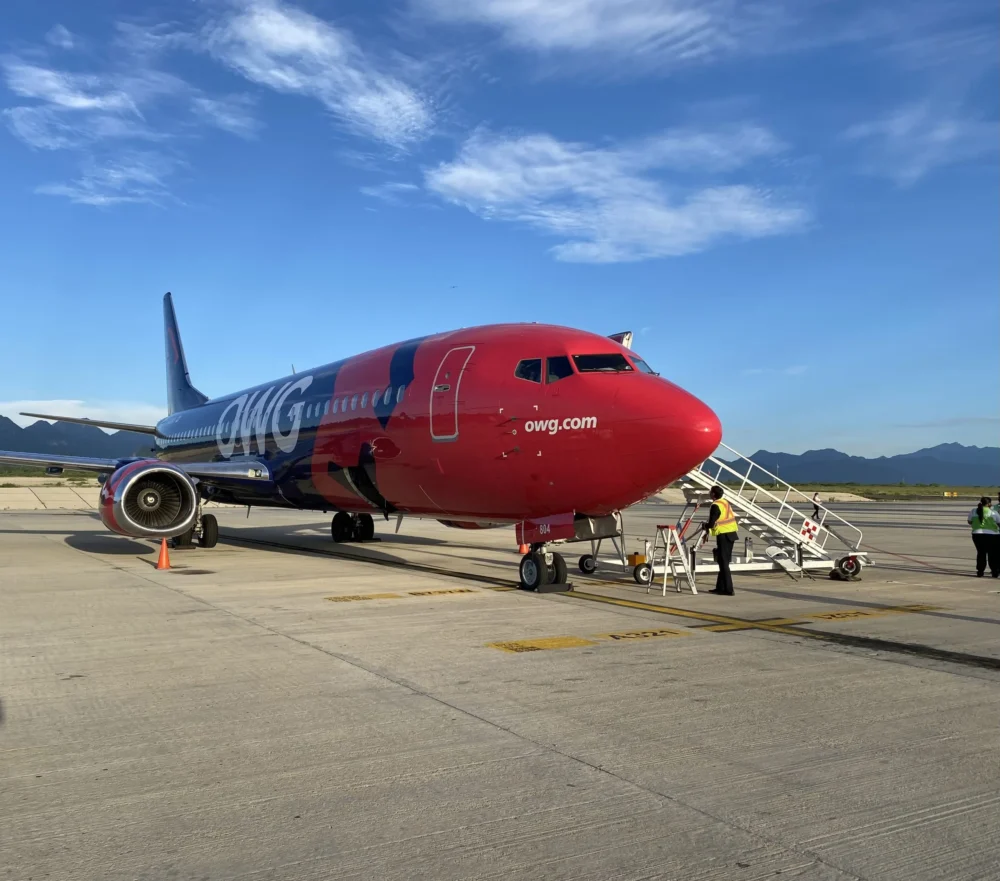 A red and blue airplane with "OWG" branding is parked on the tarmac, with ground crew preparing to assist passengers boarding via stairs.
