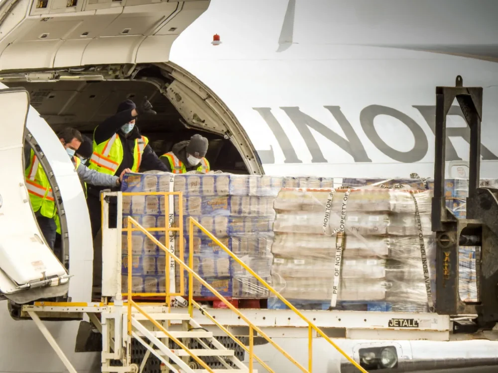 Workers in safety vests unload cargo from an airplane, stacking boxes on a loading dock. Masks are worn for safety.