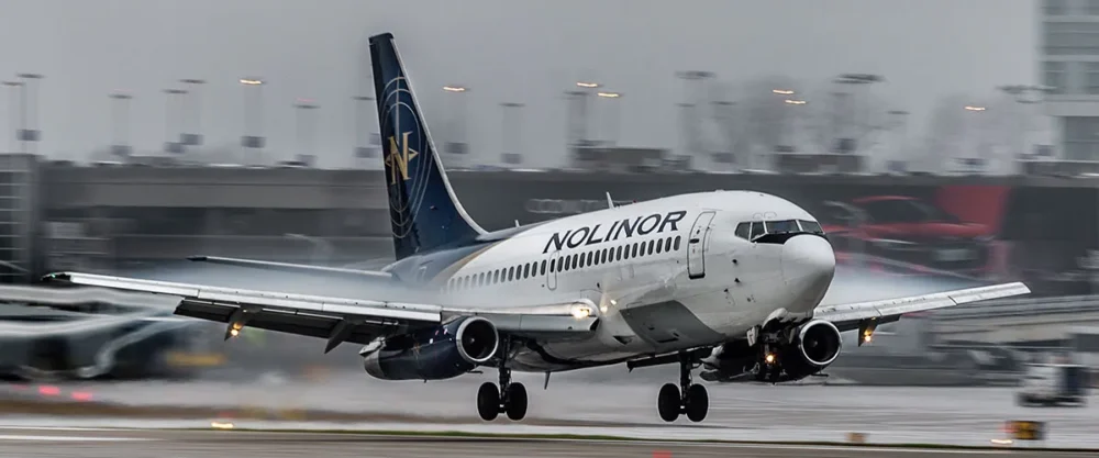 A Noliner aircraft lands on a wet runway, with rain and mist creating a dramatic backdrop at an airport.