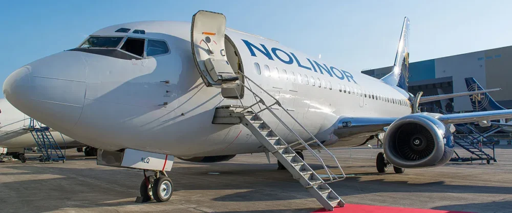A Nolinor airplane parked on the tarmac, with a boarding staircase attached and a clear blue sky in the background.