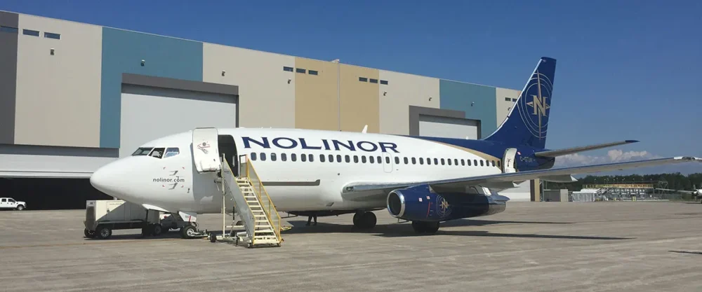 Un avion de la compagnie Nolinor est stationné sur le tarmac, avec une échelle d'embarquement à côté. Des bâtiments en arrière-plan.