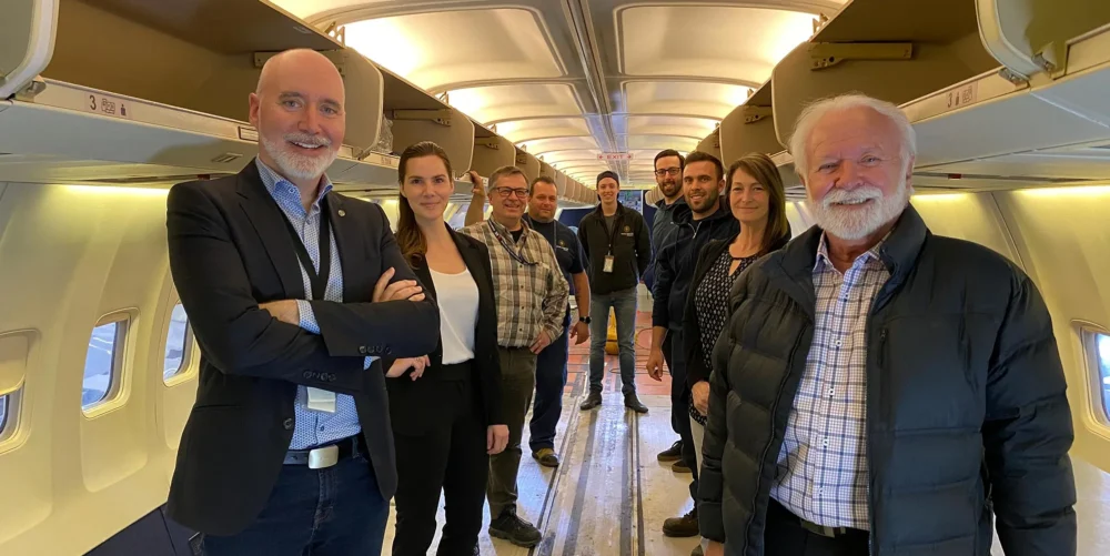 Un groupe de neuf personnes souriantes se tient dans un couloir d'avion, avec des sièges visibles de chaque côté.