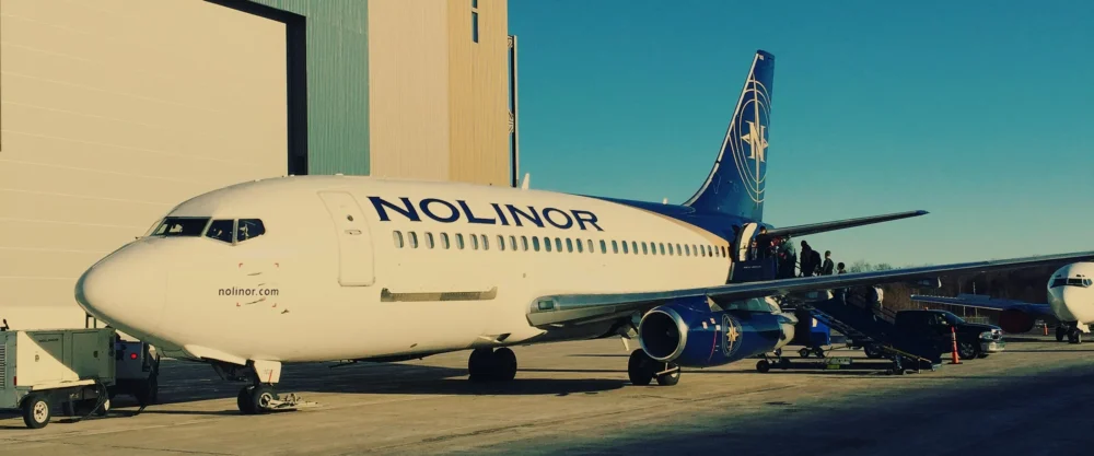 Un avion de la compagnie Nolinor est stationné sur le tarmac, des passagers montent à bord par une échelle. Le ciel est dégagé.
