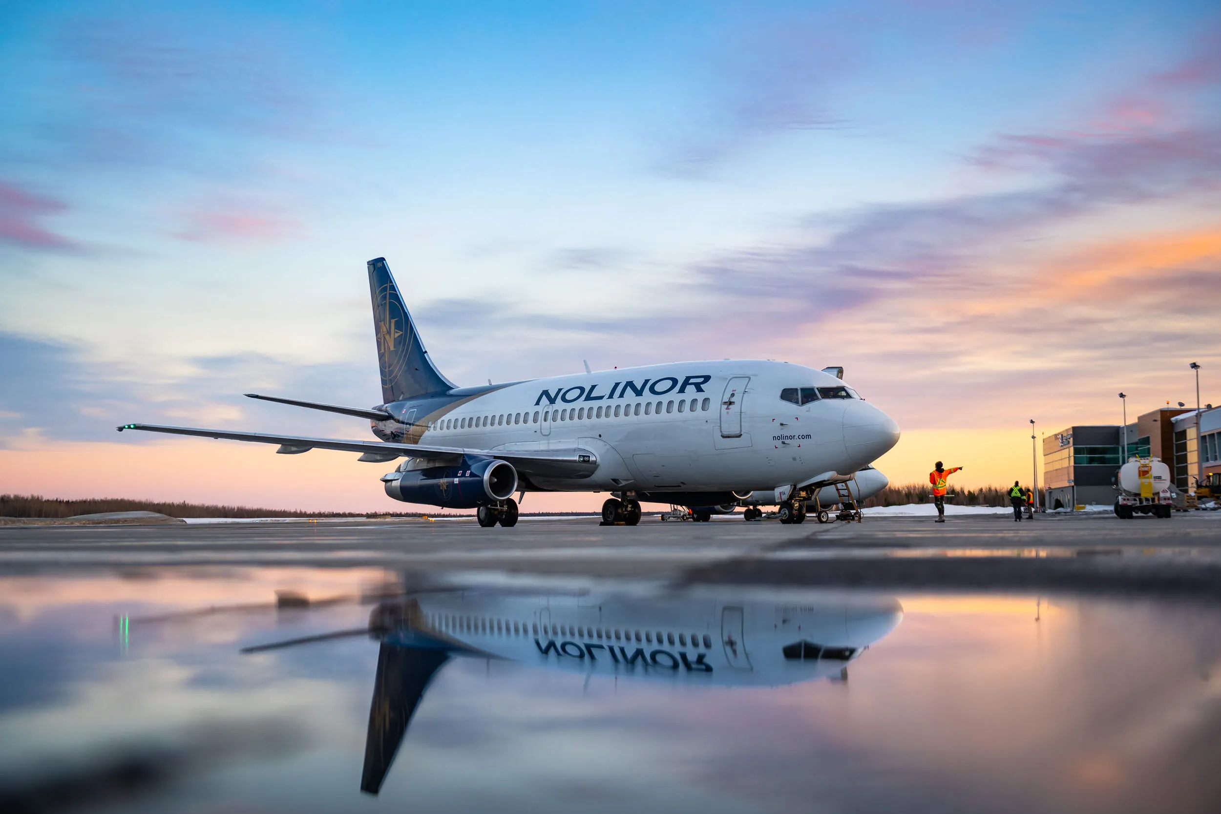 A Noliner airplane parked on a runway at sunset, reflecting in a puddle, with ground crew preparing for departure.
