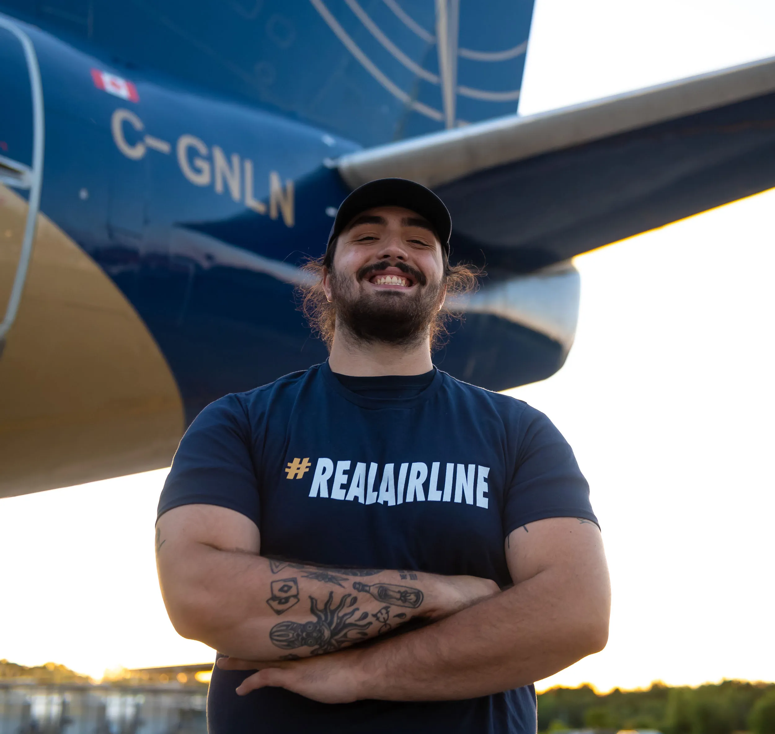 A smiling man stands with arms crossed in front of an aircraft, wearing a "#REALAIRLINE" t-shirt. The setting is outdoors during sunset.