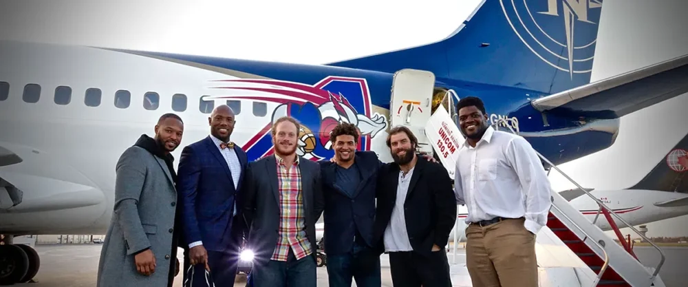 A group of six men stands in front of a blue airplane featuring a logo, smiling and posing together.