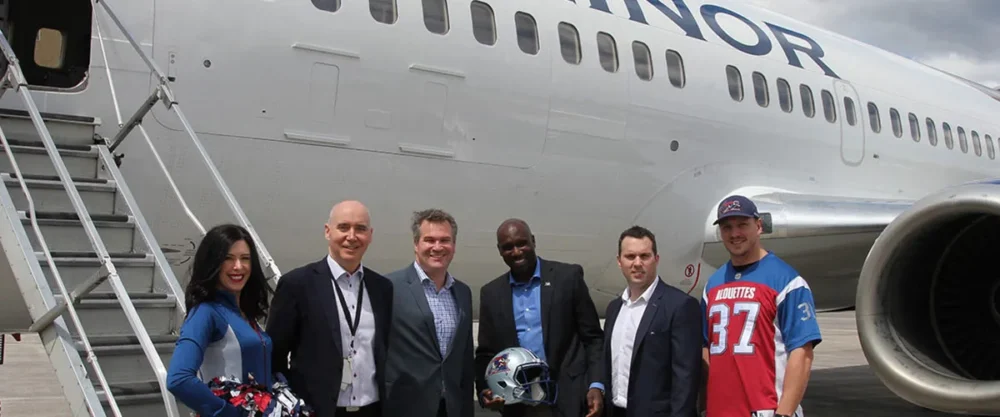 Group of six people, including cheerleaders and team members, pose with a football helmet near a plane marked "MINOR."