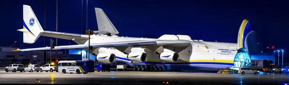 A large cargo plane is parked at night, with its cargo door open, revealing loading operations. Several vehicles are nearby.