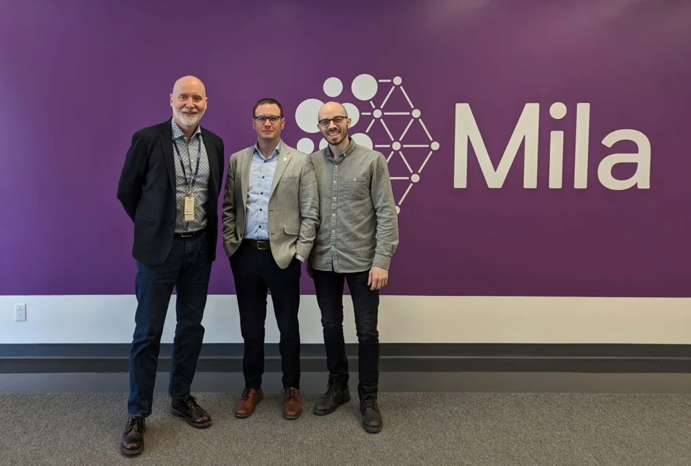 Three men stand together in front of a purple wall with the "Mila" logo, smiling and dressed in business casual attire.