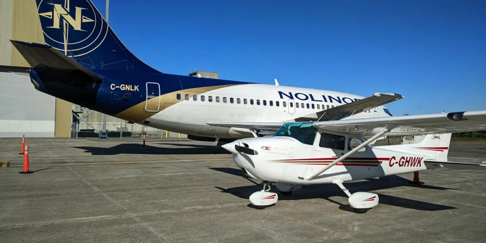 Un petit avion Cessna blanc et rouge est garé devant un grand avion commercial aux couleurs de Nolinor, sous un ciel bleu.