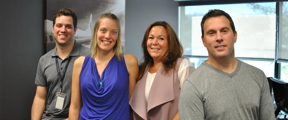 Four smiling individuals stand together in an office setting, with natural light coming through large windows.