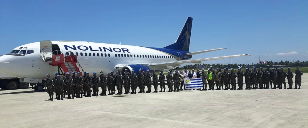 A group of soldiers stands in formation on an airstrip, with a Nolinor airplane in the background and a flag displayed.