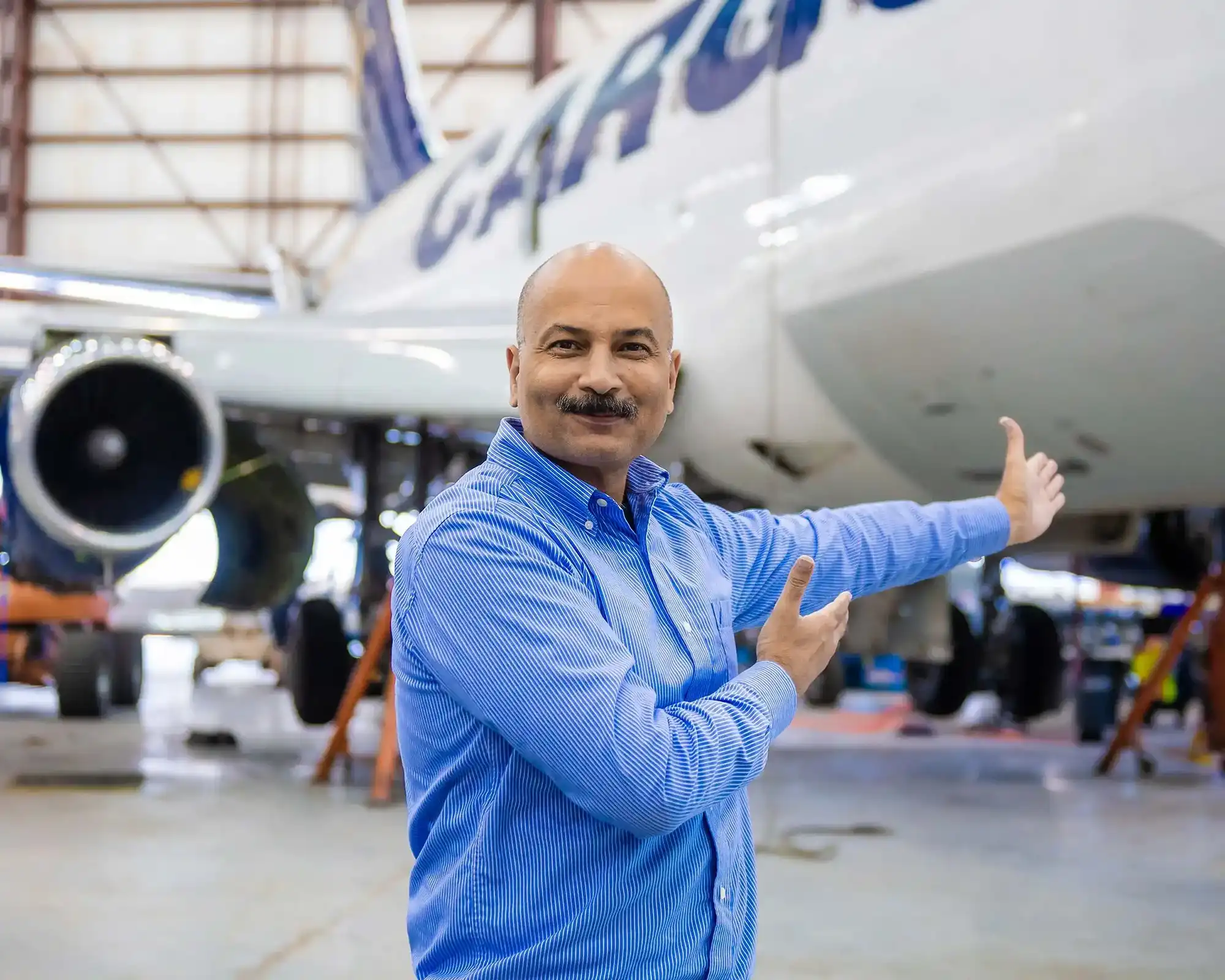 A smiling man is pointing at a transport plane in the background, inside an aircraft maintenance hangar. He is wearing a blue shirt.