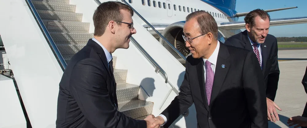 A man in a suit shakes hands with another man wearing glasses and a suit at an airport, with a plane in the background.