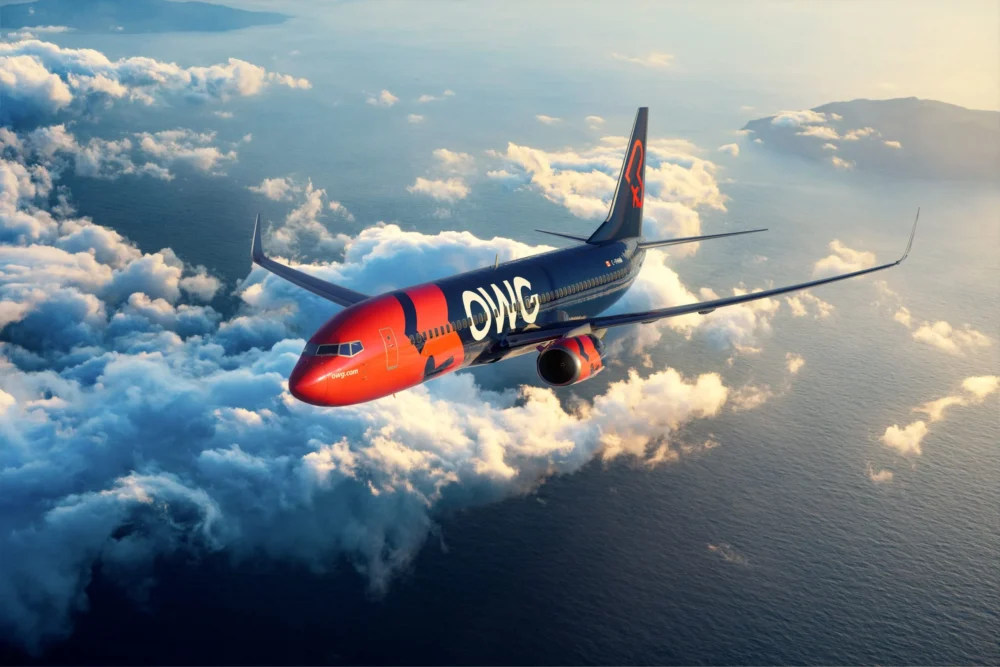 A brightly colored airplane with "OWG" on its fuselage flies above fluffy clouds over the ocean, with a distant landmass visible.