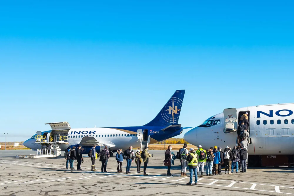 Passengers disembark from a plane while others wait near two parked aircraft under a clear blue sky.