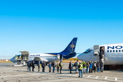 Passengers disembark from a plane while others wait near two parked aircraft under a clear blue sky.