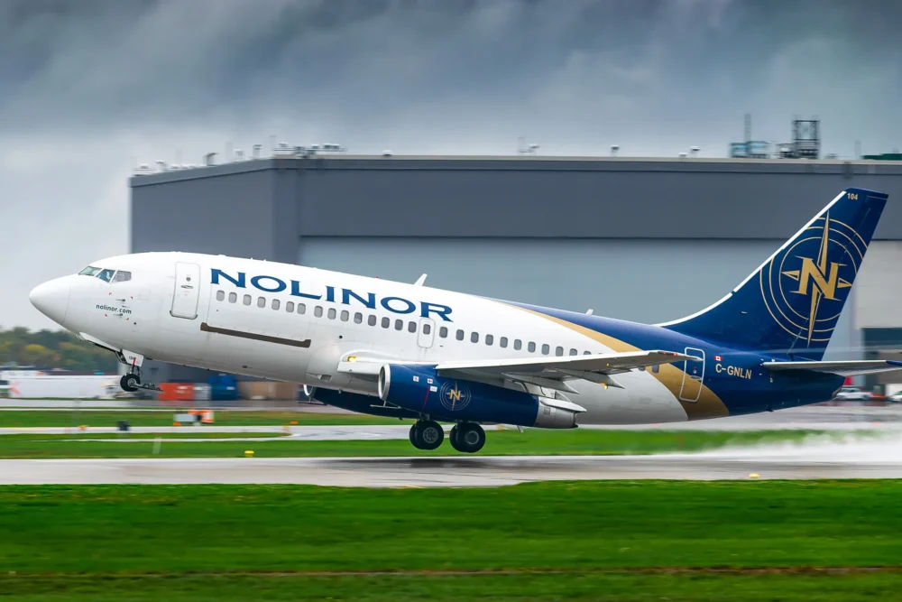 Nolinor Airlines Boeing 737 takes off from a runway, with a gray sky and industrial building in the background.