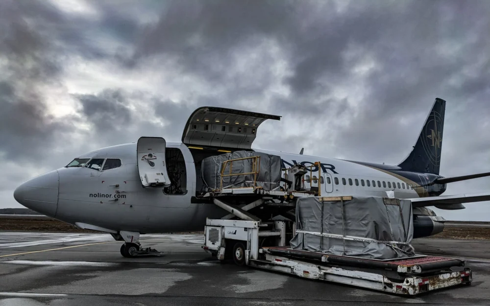 A cargo plane with its rear door open is being loaded with freight on a cloudy day at an airport.