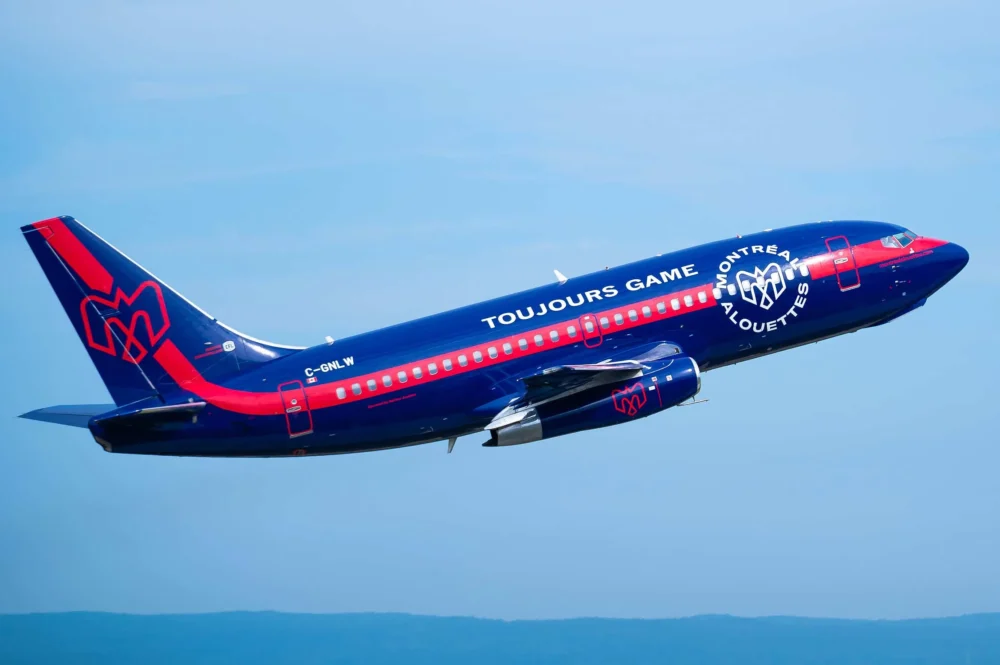 A colorful airplane with a blue and red design takes off against a clear sky, featuring the text "Toujours Game" and "Montreal Alouettes."