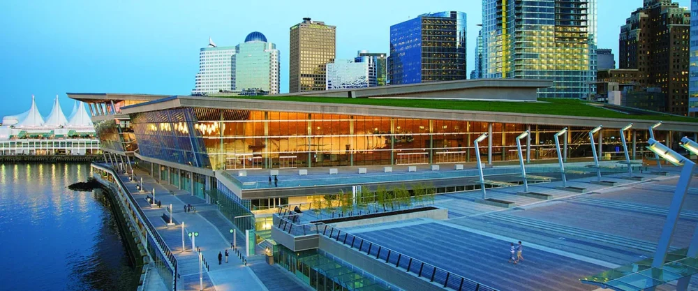 Modern waterfront building with large glass windows reflecting city lights, surrounded by a paved plaza and skyline.