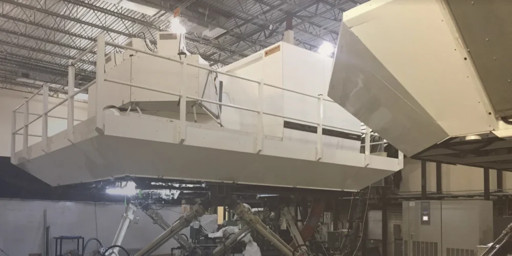 A large white structure with railings is positioned in a warehouse, connected to machinery and equipment for testing or simulation.
