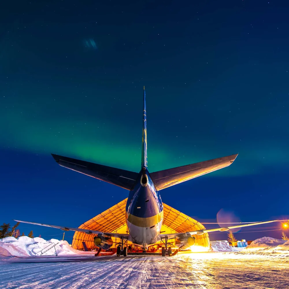 A large airplane is parked in front of a hangar under a night sky illuminated by the northern lights. Snow covers the ground.