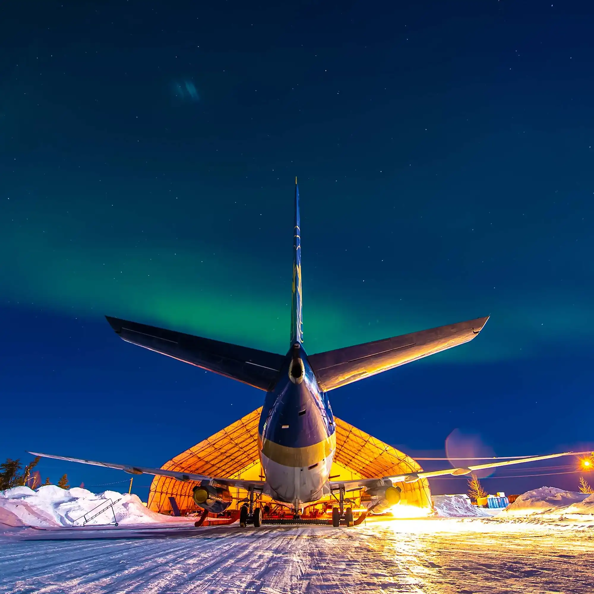 A large airplane is parked in front of a hangar under a night sky illuminated by the northern lights. Snow covers the ground.
